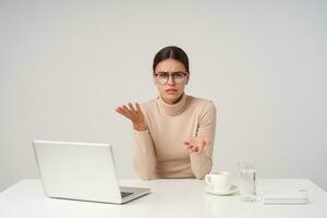 Bewildered young attractive dark haired woman dressed in beige poloneck sitting at table in modern office, having tense conversation and raising emotionally hands, isolated over white background photo