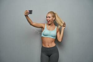 Joyful beautiful young healthy blonde female lady with ponytail hairstyle holding mobile phone in raised hand and smiling widely to camera while making selfie, isolated over grey background photo