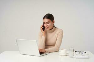 Shot of young pretty positive dark haired lady with natural makeup smiling cheerfully while making call and looking at screen while typing text on keyboard, isolated over white background photo