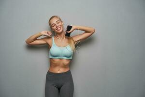 Plesant looking sleepy young slim blonde lady with casual hairstyle stretching herself before morning training, keeping eyes closed and smiling positively while posing over grey background photo