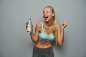 Indoor photo of charming happy sporty female with ponytail hairstyle laughing joyfully and holding bottle of water, being satisfied with evening workout, isolated over grey background