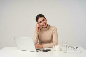 Positive young attractive brunette businesswoman with ponytail hairstyle touching her face with raised hand while sitting over white background and smiling cheerfully photo