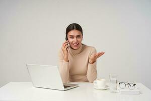 Indoor shot of excited young beautiful dark haired woman with ponytail hairstyle making call to her friend while having break with her work, smiling cheerfully at camera and keeping palm raised photo