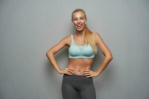 Studio photo of happy young slim female with long blonde hair keeping hands on her waist and looking at camera joyfully, wearing mint sporty top and grey leggins over grey background