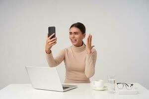 Studio shot of young attractive dark haired lady raising hand with smartphone while having exciting phone conversation, working at office with modern laptop photo