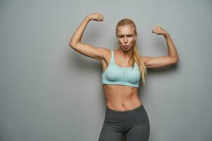 Indoor photo of cheerful young slim blonde female with casual hairstyle demonstrating her power in raised hands, grimacing to camera while posing over grey background