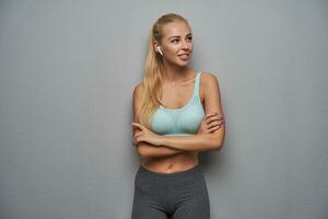 Attractive young slim woman with long blonde hair standing over grey background with folded hands on her chest, looking aside with charming smile and wearing earpieces photo