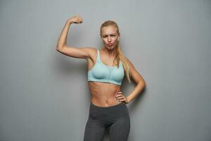 Portrait of funny young sporty blonde woman raising hand and showing her power, frowning eyebrows and pouting lips while posing over grey background, dressed in athletic clothes photo