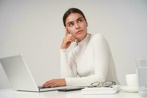 Beautiful young brunette female with ponytail hairstyle working with her laptop at office, holding her head with raised hand and looking pensively aside, isolated over white background photo