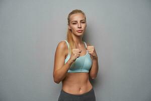 Serious sporty young long haired blonde female in athletic wear standing over light grey background photo