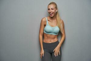 Overjoyed young sporty blonde model with casual hairstyle looking aside and laughing happily, wearing mint top and grey leggins while posing over grey background with hands down photo