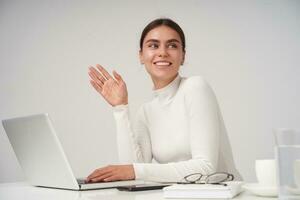 Cheerful young beautiful dark haired lady with natural makeup raising hand in hello gesture and looking positively aside with friendly smile, isolated over white background photo
