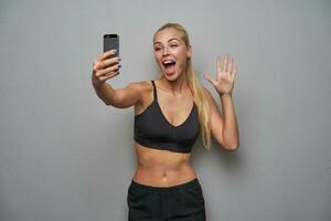 Indoor shot of joyful young sporty blonde female with casual hairstyle holding mobile phone photo