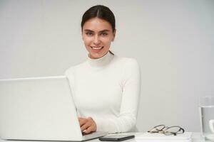 interior Disparo de alegre bonito joven morena dama con natural maquillaje acuerdo manos en teclado mientras mecanografía texto y sonriente felizmente a cámara, aislado terminado blanco antecedentes foto