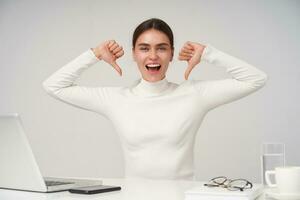 Joyful young attractive brunette female with natural makeup pointing happily to herself with raised hands and looking at camera with excited face, sitting at table over white background photo