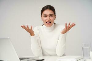 Irritated young pretty dark haired female with natural makeup raising emotionally her hands while looking excitedly at camera, dressed in formal clothes while posing over white background photo