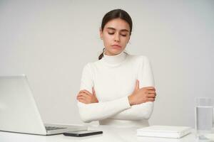 Upset young beautiful dark haired female with ponytail hairstyle folding hands on her chest and looking sadly down while sitting over white background with modern laptop photo