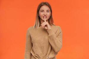 Studio photo of young blonde woman with short haircut keeping forefinger on her lips while asking to keep her secret and smiling cunningly, isolated over orange background