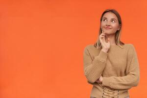 Pleased young attractive blonde woman with short haircut smiling gently while looking upwards and touching face with raised hand, isolated over orange background photo