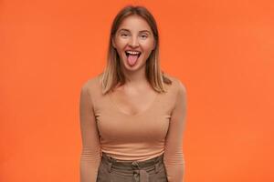 Joyful young attractive white-headed woman with short haircut showing cheerfully tongue while fooling and keeping hands down while standing over orange background photo