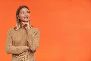 Indoor photo of young lovely white-headed woman with casual hairstyle biting her underlip while looking positively aside and leaning chin on raised hand, isolated over orange background