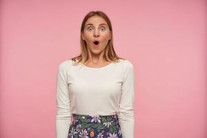 Suprised pretty young female with blonde hair wearing white blouse and flowered skirt while standing over pink background, rounding eyes and raising eyebrows amazedly while looking to camera photo