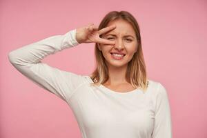 Indoor photo of young blonde female in casual wear raising hand with victory sign to her face, showing her white perfect teeth and frowning eyebrows over pink background