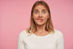 Shocked attractive young blonde woman biting underlip and looking at camera with wide eyes opened, raising eyebrows and wrinkling forehead over pink background photo