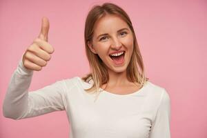 Indoor shot of happy beautiful young blonde woman with casual hairstyle looking at camera with wide mouth opened and showing raised thumb, isolated over pink background photo