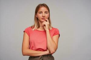 Studio photo of thoughtful pretty young lady with short blonde hair wearing casual hairstyle while posing over white background, holding chin with hand and looking at camera with folded lips