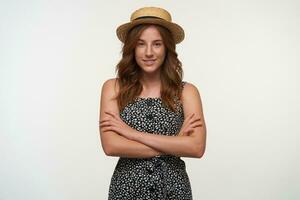 Portrait of attractive young woman with foxy wavy hair standing over white background with folded hands, looking to camera with charming smile, wearing romantic dress and boater hat photo