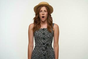 Portrait of pretty confused young redhead female with hands down wearing romantic dress and boater hat, looking at camera with wide mouth opened and frowning, standing over white background photo