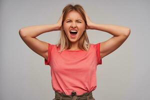 Stressed young pretty blonde female with casual hairstyle holding her head with raised hands, frowning her face with wide mouth opened while looking at camera, isolated over white background photo