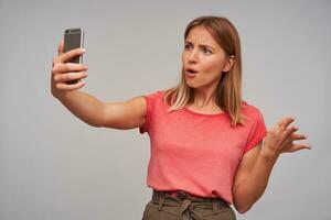 Indoor shot of confused young blonde female in casual clothes wearing natural makeup while having phone conversation, standing over white background with smartphone in raised hand photo