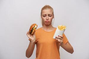 Upset young pretty blonde woman in orange t-shirt keeping unhealthy food in her hands and looking sadly at it, frowning eyebrows and twisting her mouth while posing over white background photo