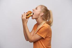 espalda ver de hambriento joven largo peludo rubia dama con casual peinado comiendo ávidamente grande Fresco hamburguesa mientras en pie en contra blanco antecedentes y acuerdo su ojos cerrado foto