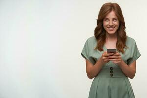 Happy attractive female with red hair posing over white background with mobile phone in hands, looking aside and smiling happily, wearing romantic dress photo