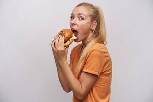 estudio foto de bonito joven largo peludo rubia mujer en casual vestir participación sabroso hamburguesa en su manos y yendo a comer él, en pie en contra blanco antecedentes