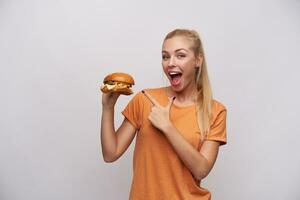 Excited young long haired blonde lady in casual clothes pointing joyfully with forefinger on big fresh burger in raised hand, looking at camera with wide happy smile while posing over white background photo