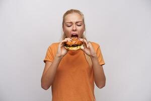 Indoor photo of young hungry long haired blonde female frowning her face while eating insatiably her tasty hamburger, dressed in orange t-shirt while standing over white background