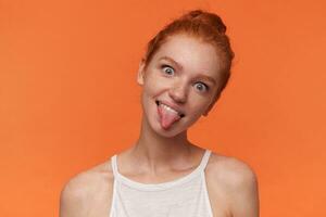 Indoor shot of ridiculous cute young female wearing her red hair in knot isolated over orange background, looking at camera with cheerful smile and showing tongue, making fun in casual clothes photo