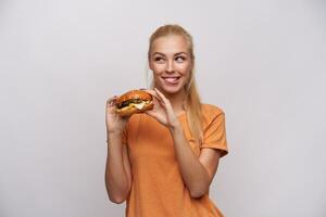 alegre joven hermosa rubia mujer con casual peinado participación hamburguesa con queso en elevado manos y mirando astutamente aparte con ancho sonrisa, aislado terminado blanco antecedentes foto