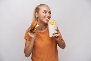 Indoor photo of attractive young blonde lady in orange t-shirt smiling cheerfully while pulling with teeth french fries and keeping hamburger in other hand, isolated over white background