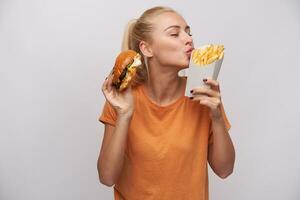 Pleased good looking young lovely blonde female in casual clothes standing over white background with fast food in raised hands, keeping eyes closed while foretasting yummy meal photo