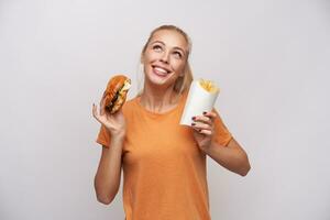 Cheerful attractive young blue-eyed blonde female holding burger and french fries in raised hands and looking happily upwards, smiling widely while posing over white background photo