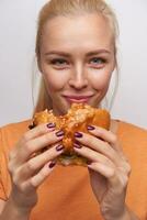 de cerca de hermosa de ojos azules joven rubia mujer comiendo rápido comida y mirando alegremente a cámara, sonriente agradablemente mientras posando en contra blanco antecedentes en casual ropa foto