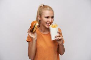 retrato de encantador joven rubia mujer con cola de caballo peinado comiendo francés papas fritas y mirando afirmativamente aparte, sonriente extensamente mientras en pie terminado blanco antecedentes foto