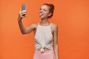 Image of attractive young lady wearing her foxy hair in knot, keeping smartphone in hand and looking at camera with funny face, fooling to camera and showing tongue, isolated over orange background photo