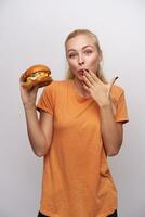 Cheerful young pretty blonde woman with casual hairstyle looking positively at camera and keeping hand on her mouth while tasting big fresh burger, standing against white background photo