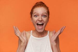 Indoor photo of joyful young readhead female with bun hairstyle wearing white top, looking to camera with surprised face and wide mouth opened, posing over orange background with raised palms